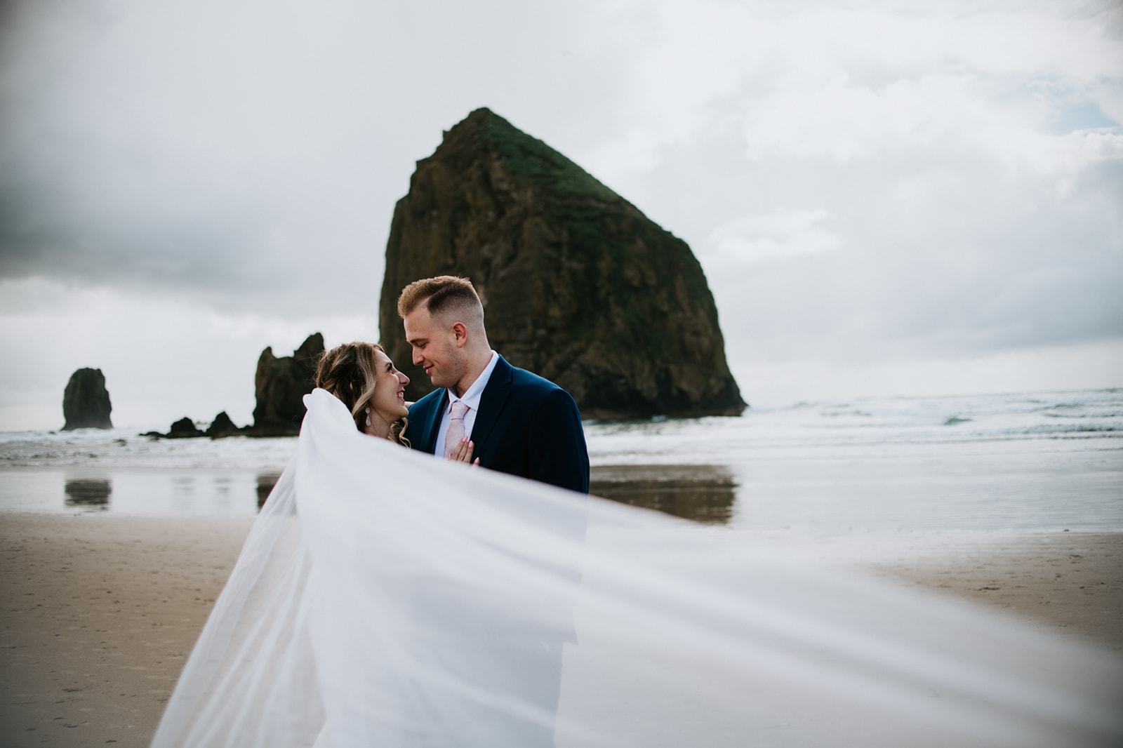 Bride looks up at her groom smiling while her veil blows in the wind with Haystack Rock behind them. 