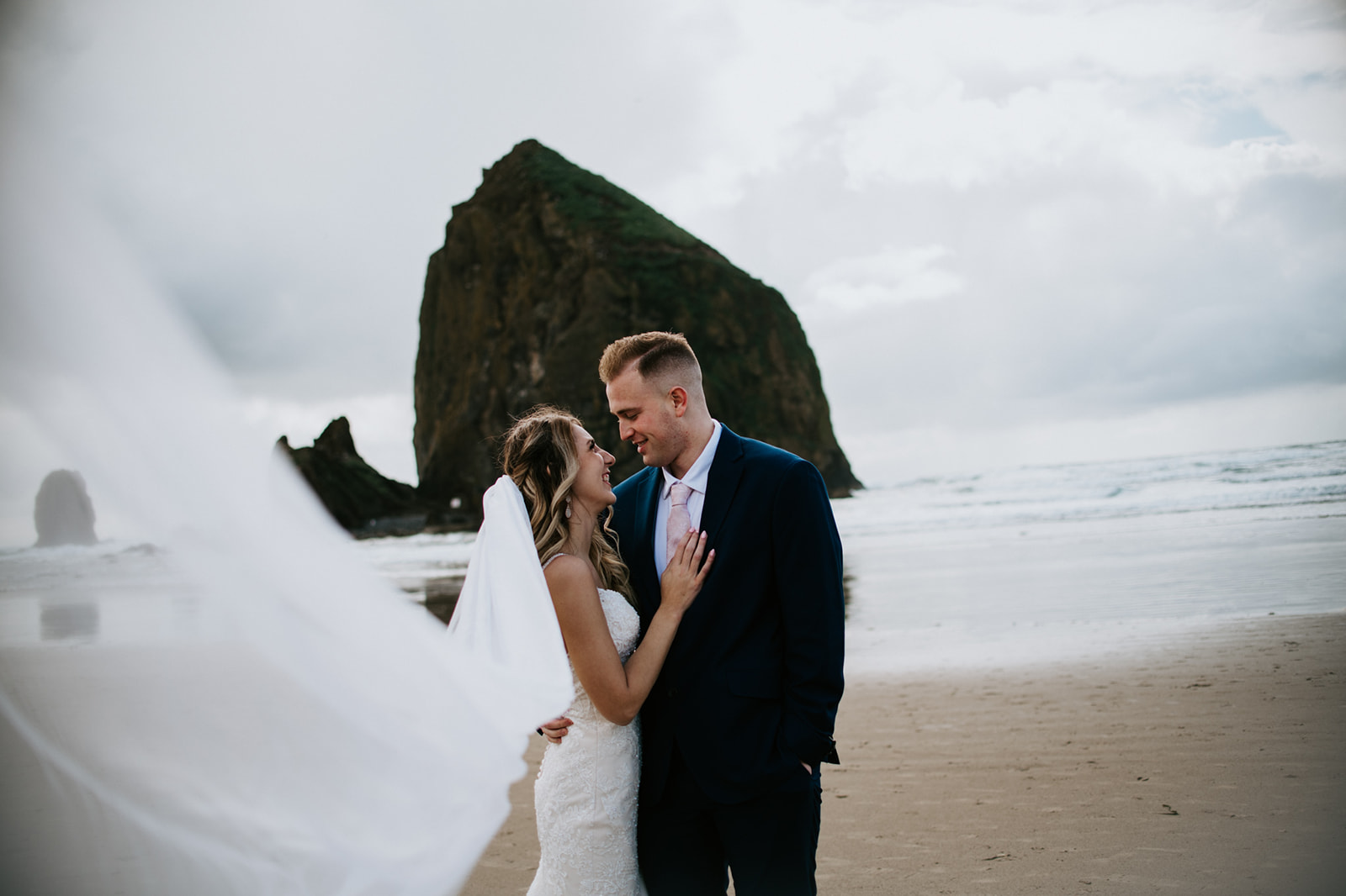 Bride's veil blows in the wind as groom looks down at her smiling. 