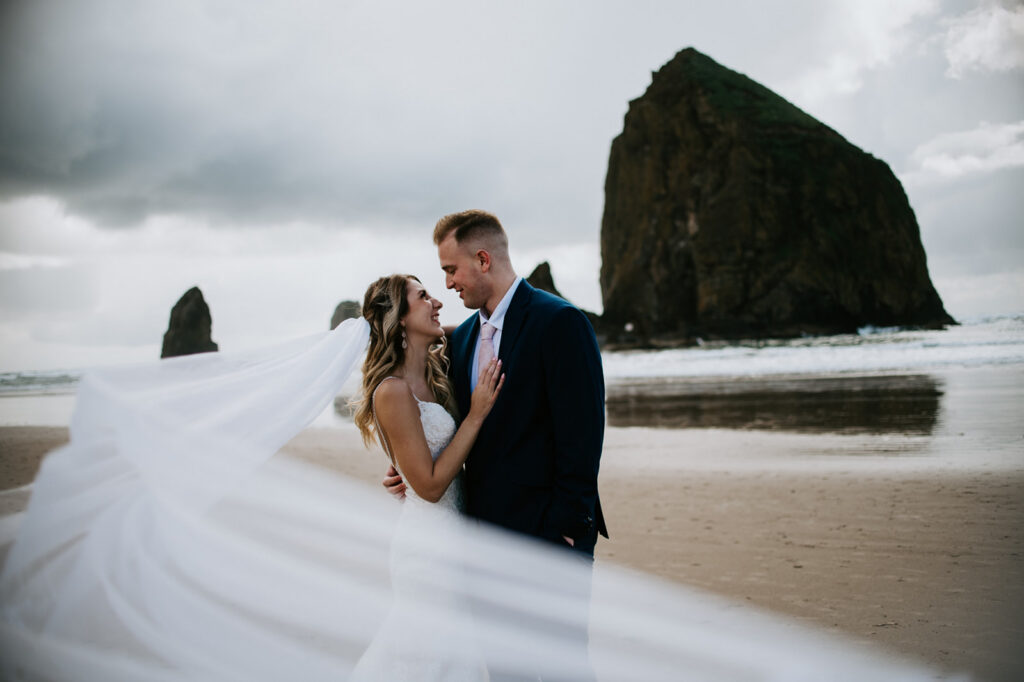 Bride and groom embracing during a windy Oregon elopement at Cannon Beach with Haystack Rock in the background and her veil blowing dramatically across the frame.