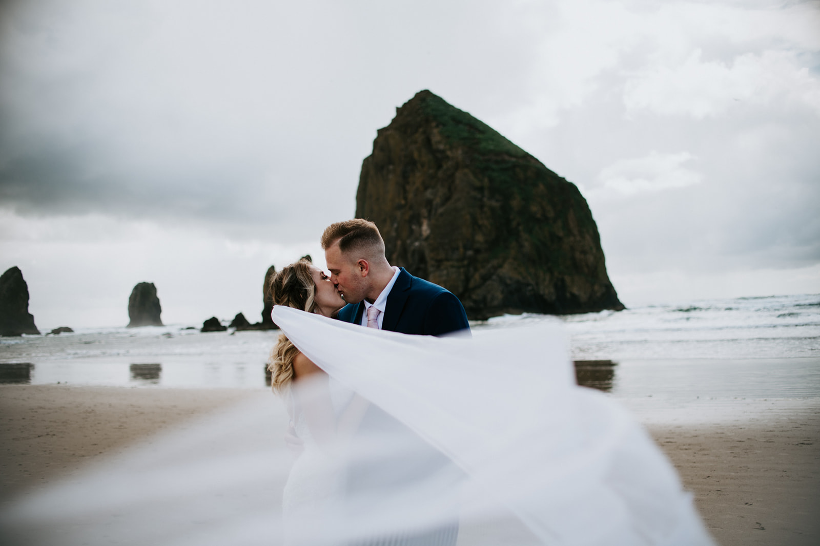 Bride and groom kiss as the bride's veil blows in the wind on Cannon Beach in front of Haystack Rock. 