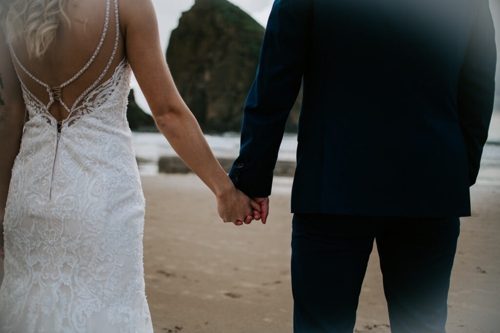 Close-up of bride and groom holding hands on the sandy beach with Haystack Rock blurred in the distance.