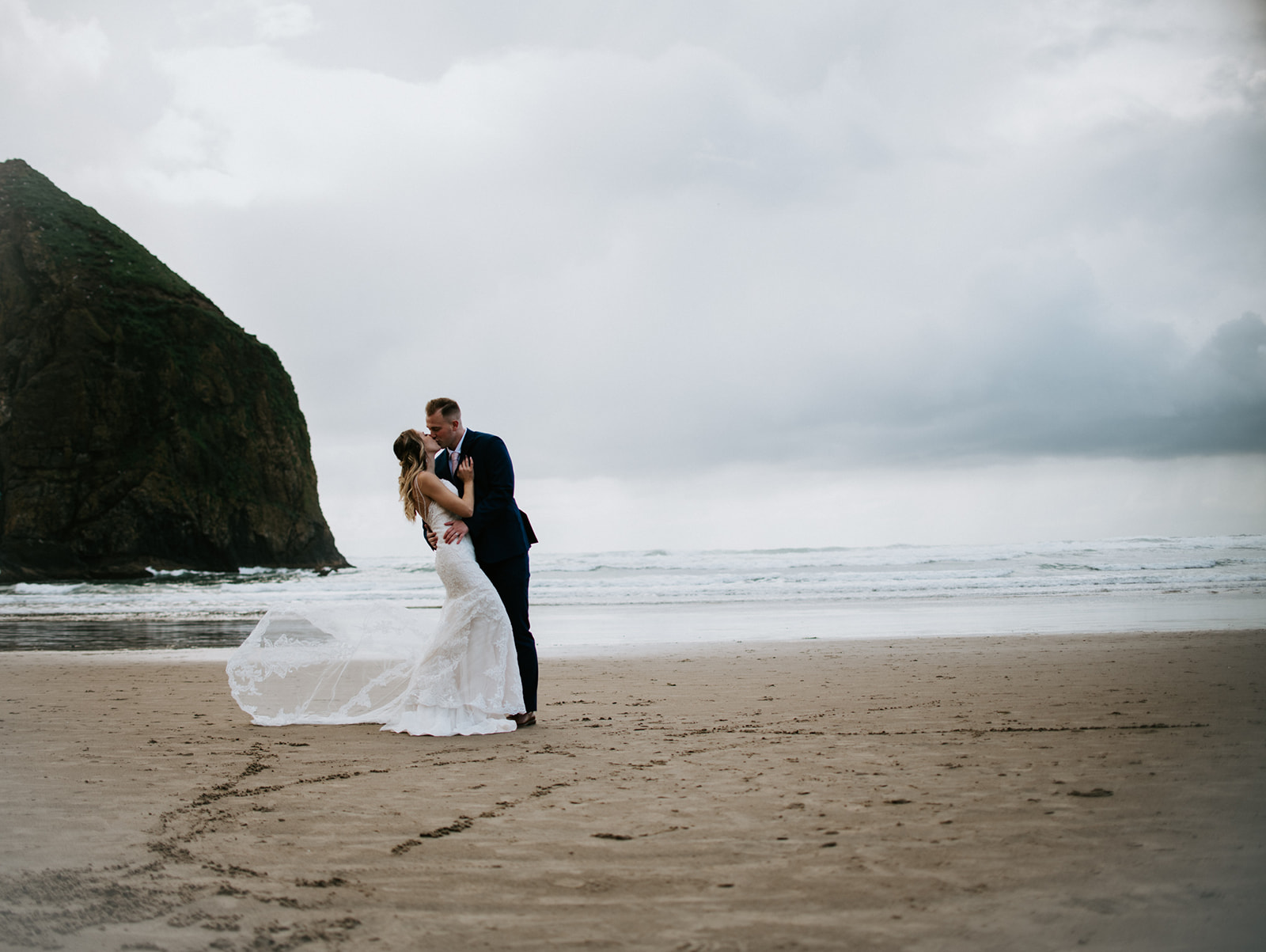 Groom begins to dip kiss the bride in front of the ocean on Cannon Beach with Haystack Rock in the background. 