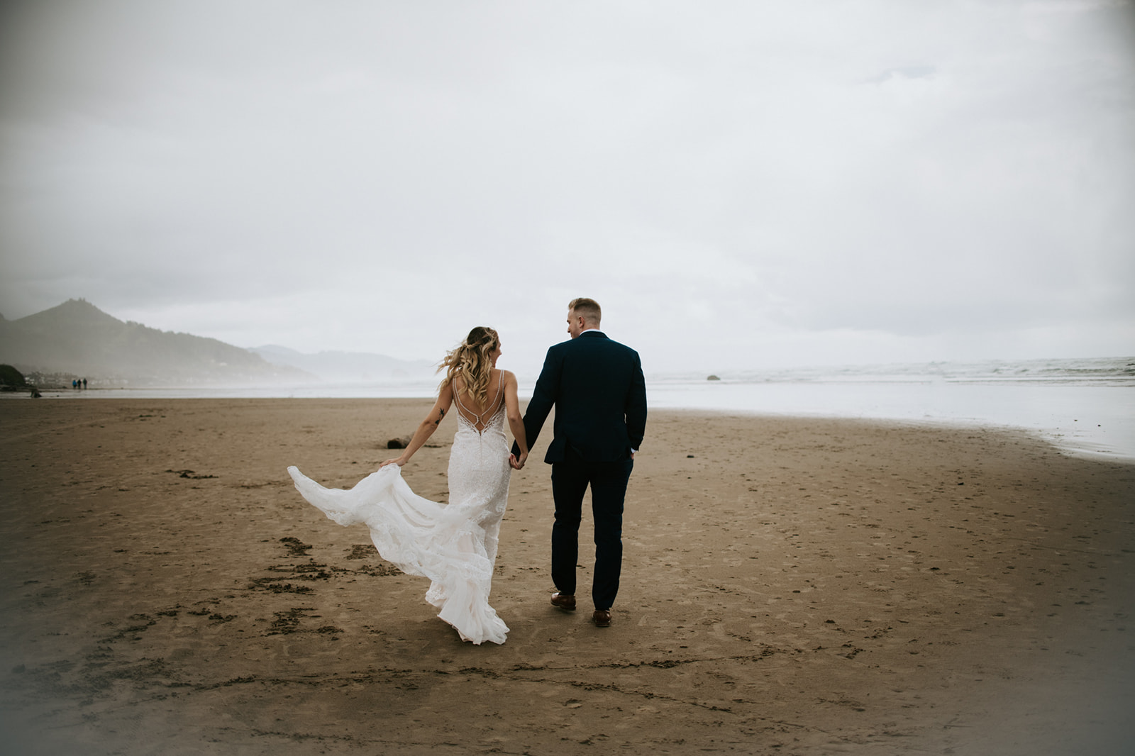 Newly married couple walks hand in hand on Cannon Beach as wind blows bride's hair and dress. 
