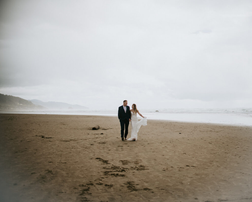 Couple walking hand in hand across a foggy Cannon Beach shoreline during their Oregon elopement with soft waves and misty coastline behind them.