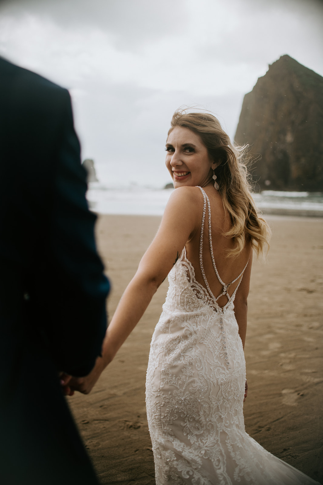 Bride smiles at the camera over her shoulder as she holds her groom's hand on Cannon Beach. 