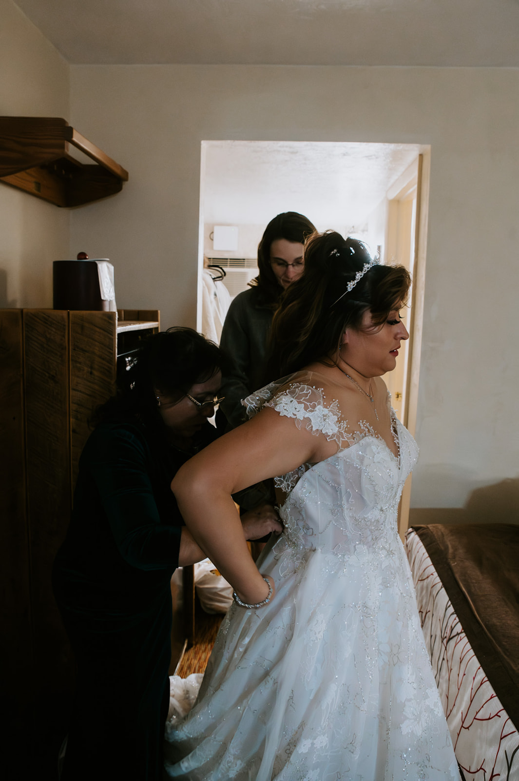 Bride’s family helping button the back of her lace wedding dress in a cozy hotel room before the ceremony.