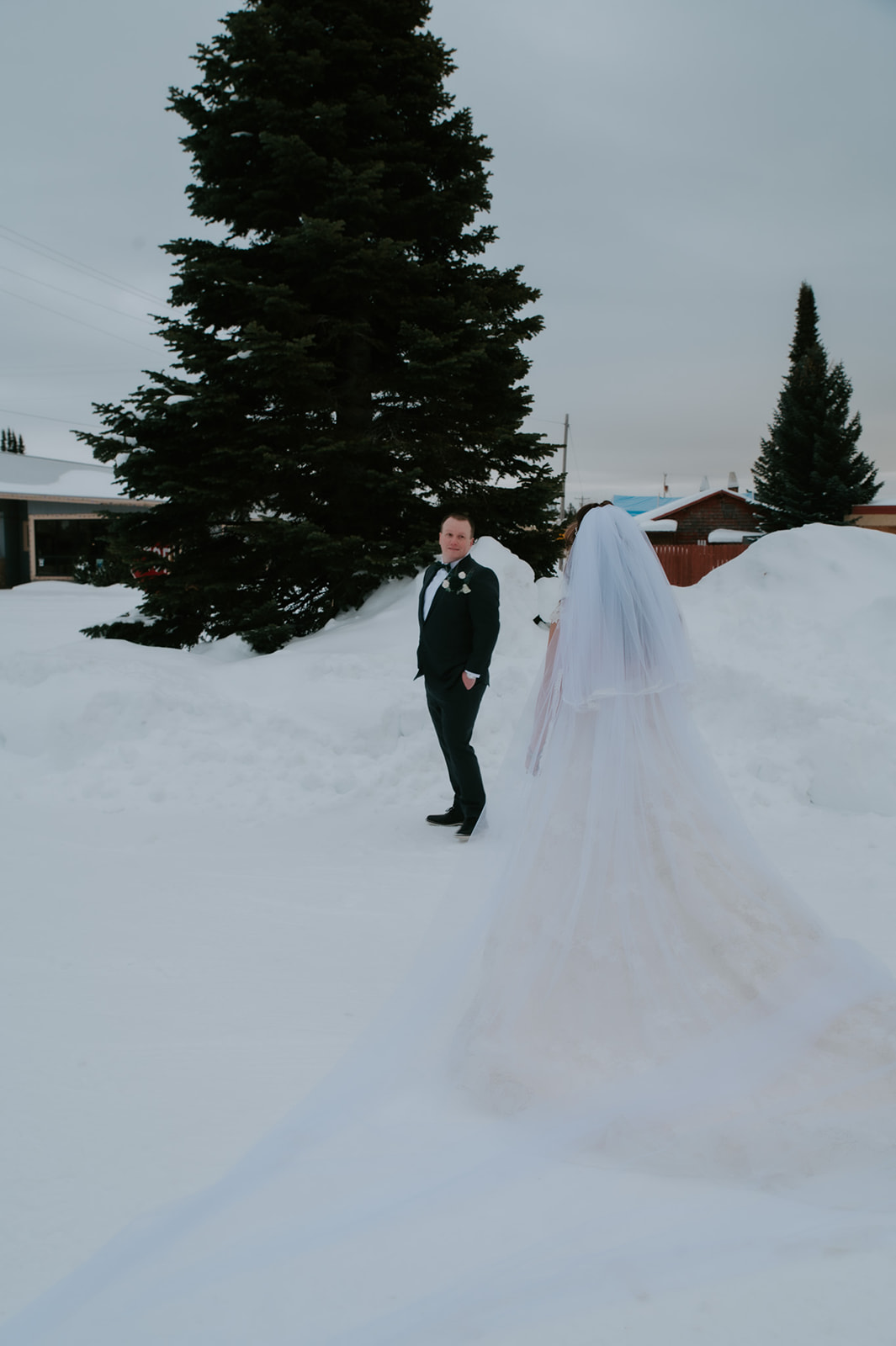 Groom waiting in the snow for their first look while the bride approaches with her long veil trailing behind her.