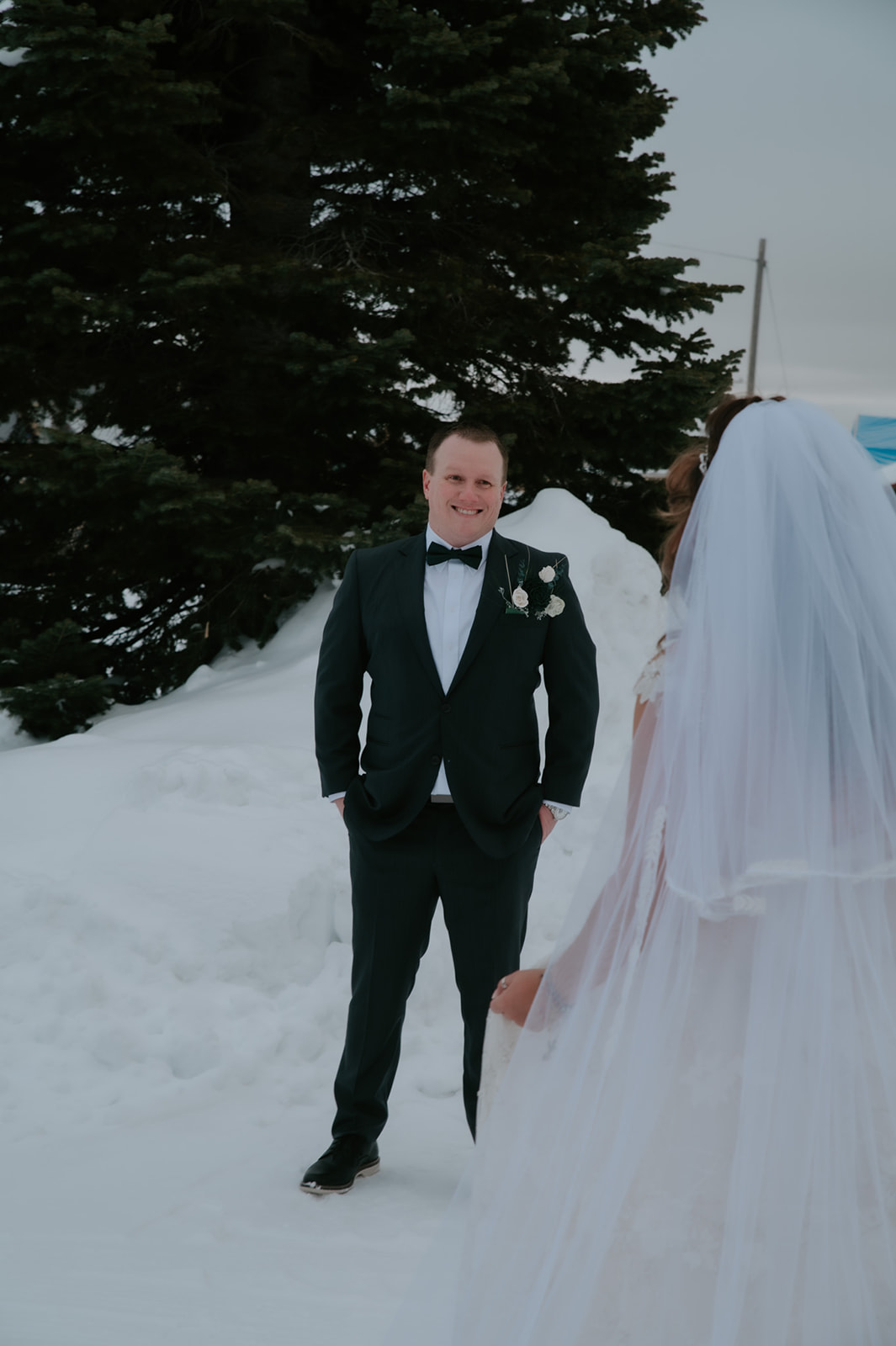 Groom smiling during a snowy first look as he sees his bride approaching through the winter landscape.