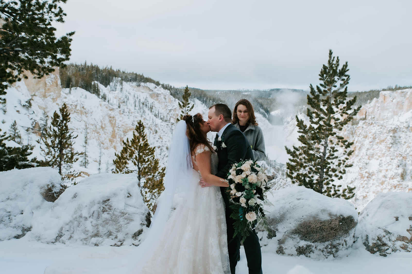 Bride and groom kissing at a snowy canyon overlook as their officiant stands nearby during the ceremony.