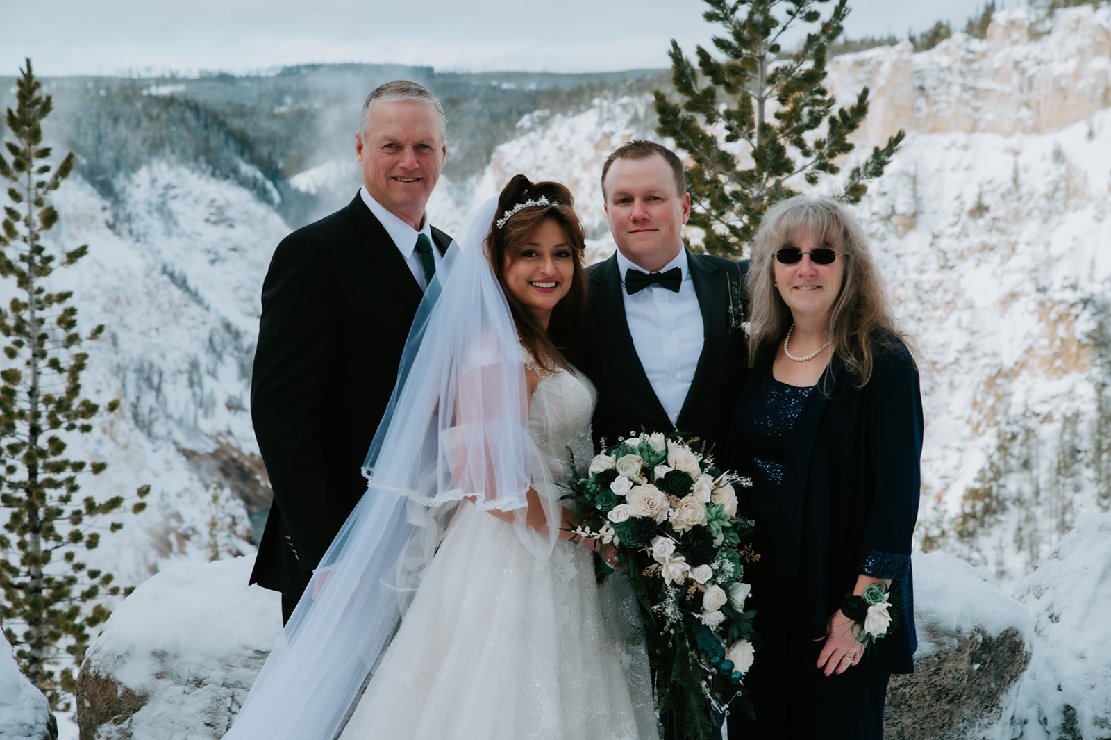 Bride and groom posing with parents at a snowy canyon overlook during their winter elopement video in Yellowstone.