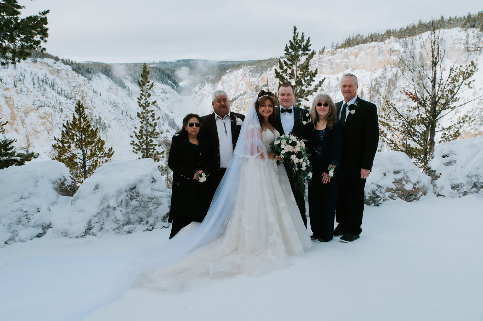 Newlyweds standing with family in front of a snow-covered canyon, bride’s long veil trailing across the ground.