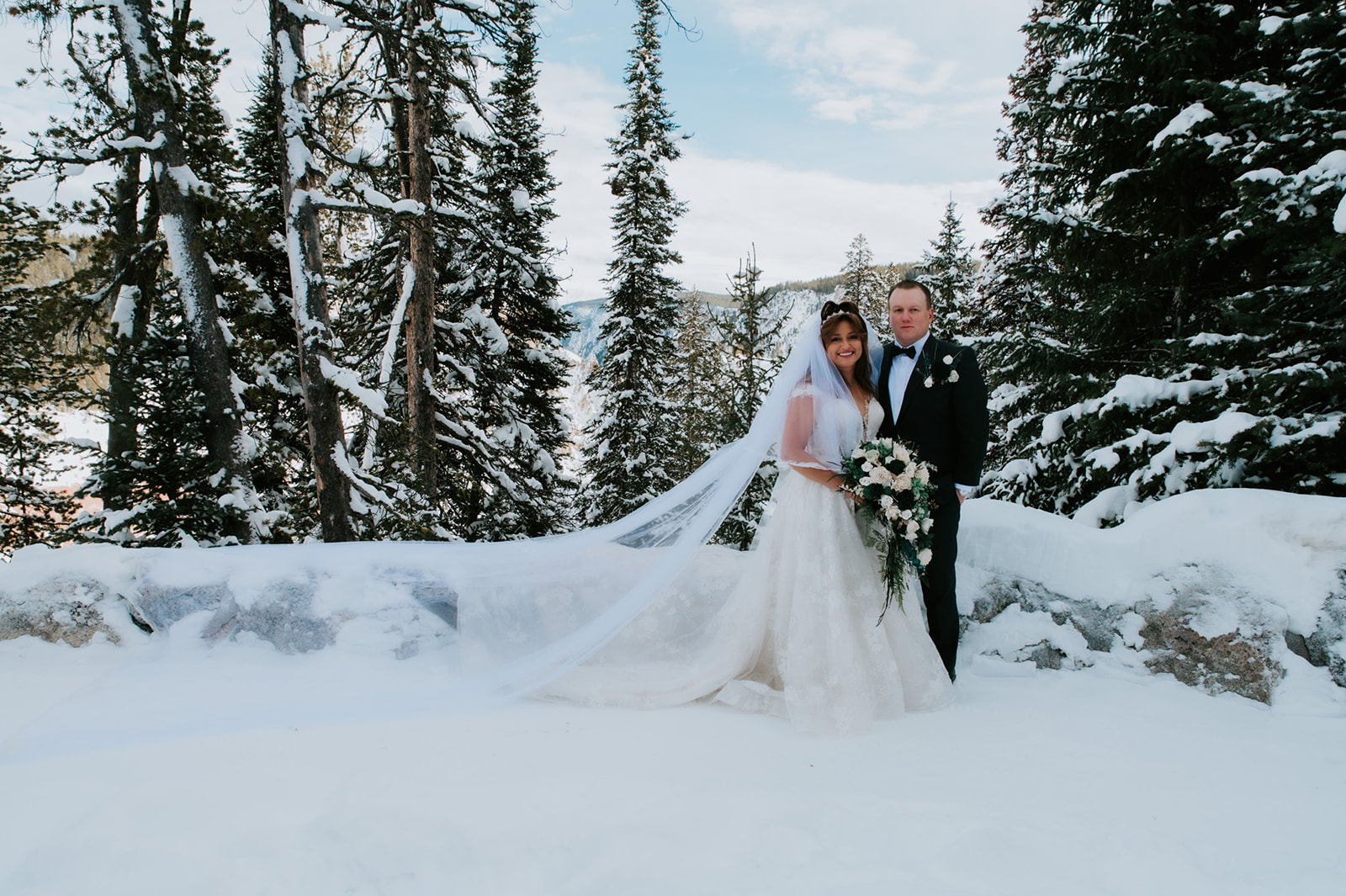 Bride and groom standing in fresh snow surrounded by tall pine trees, her cathedral veil stretched dramatically across the landscape.