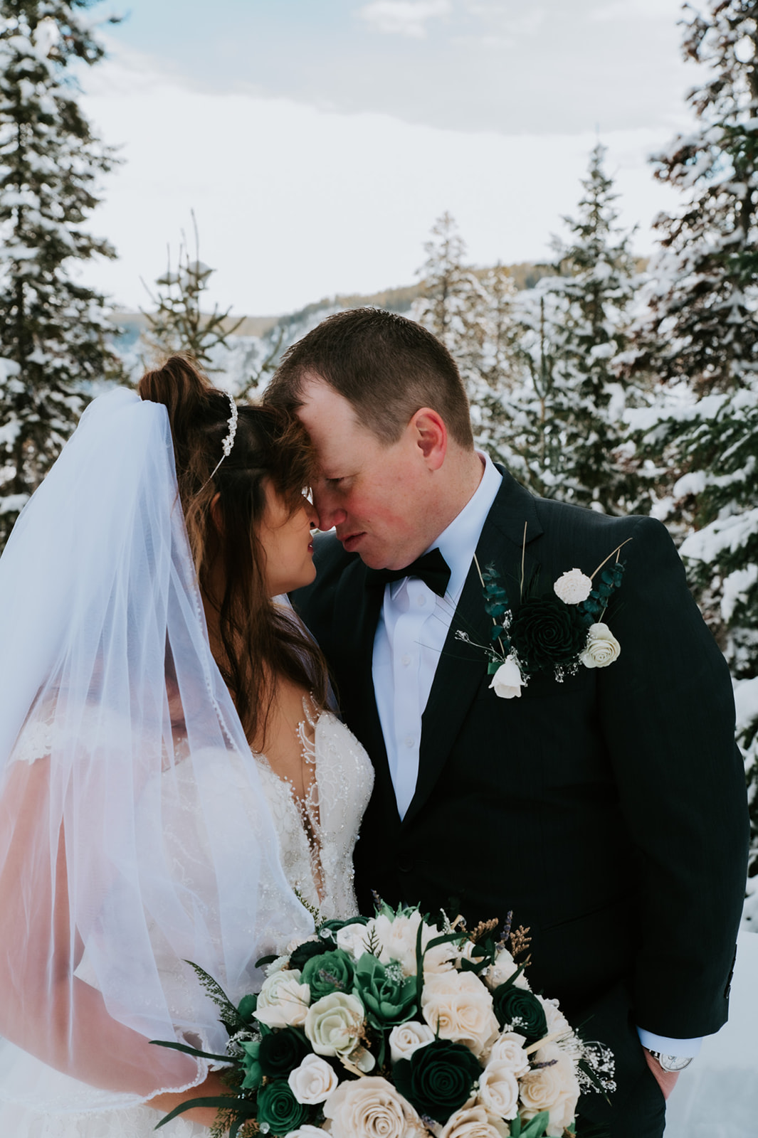 Bride and groom touching foreheads in the snow, her bouquet of white and deep green roses held between them during their intimate winter elopement video.