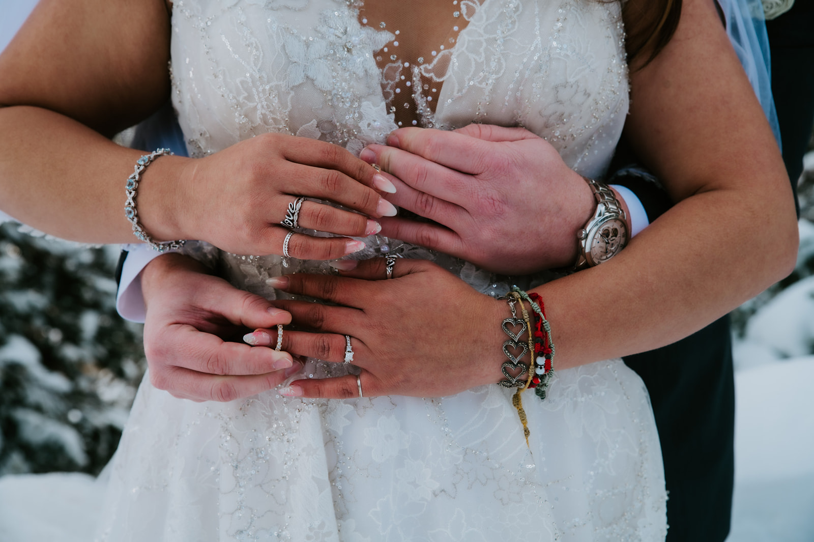 Close-up of bride and groom’s hands layered together over her lace wedding dress, highlighting rings and bracelets during their elopement video.