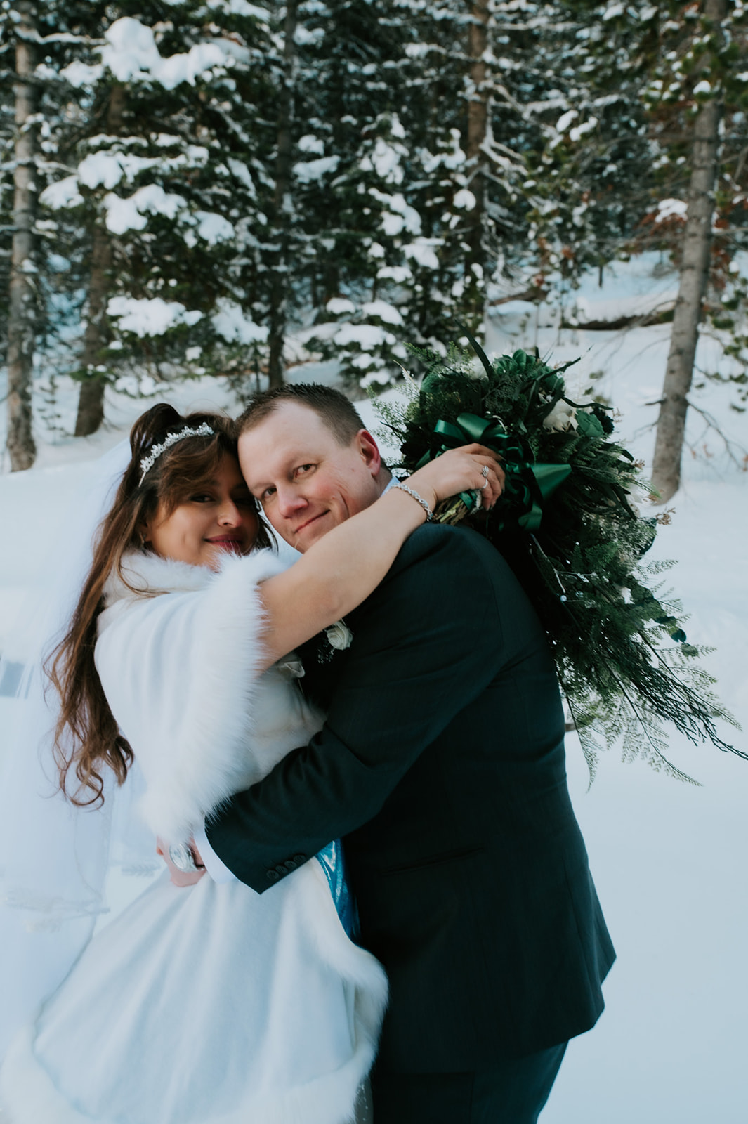 Bride and groom embracing in a snowy pine forest, her bouquet wrapped around his shoulder during their romantic elopement video.