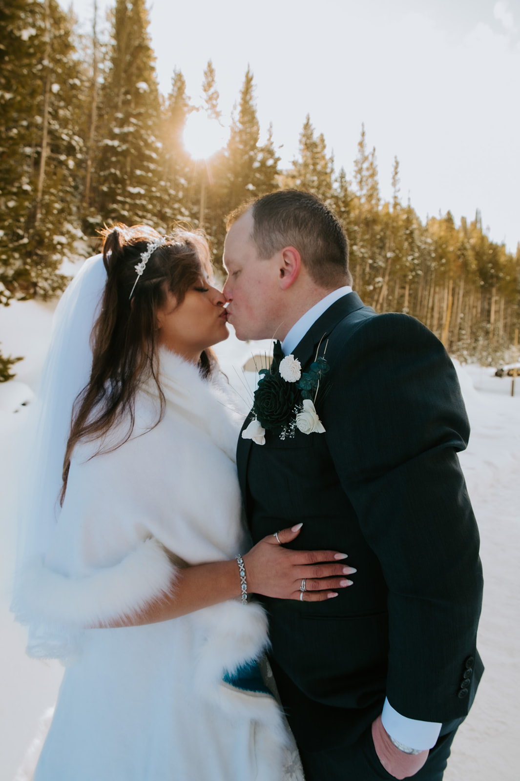 Bride and groom sharing a kiss in golden winter light with snow-covered pine trees behind them.
