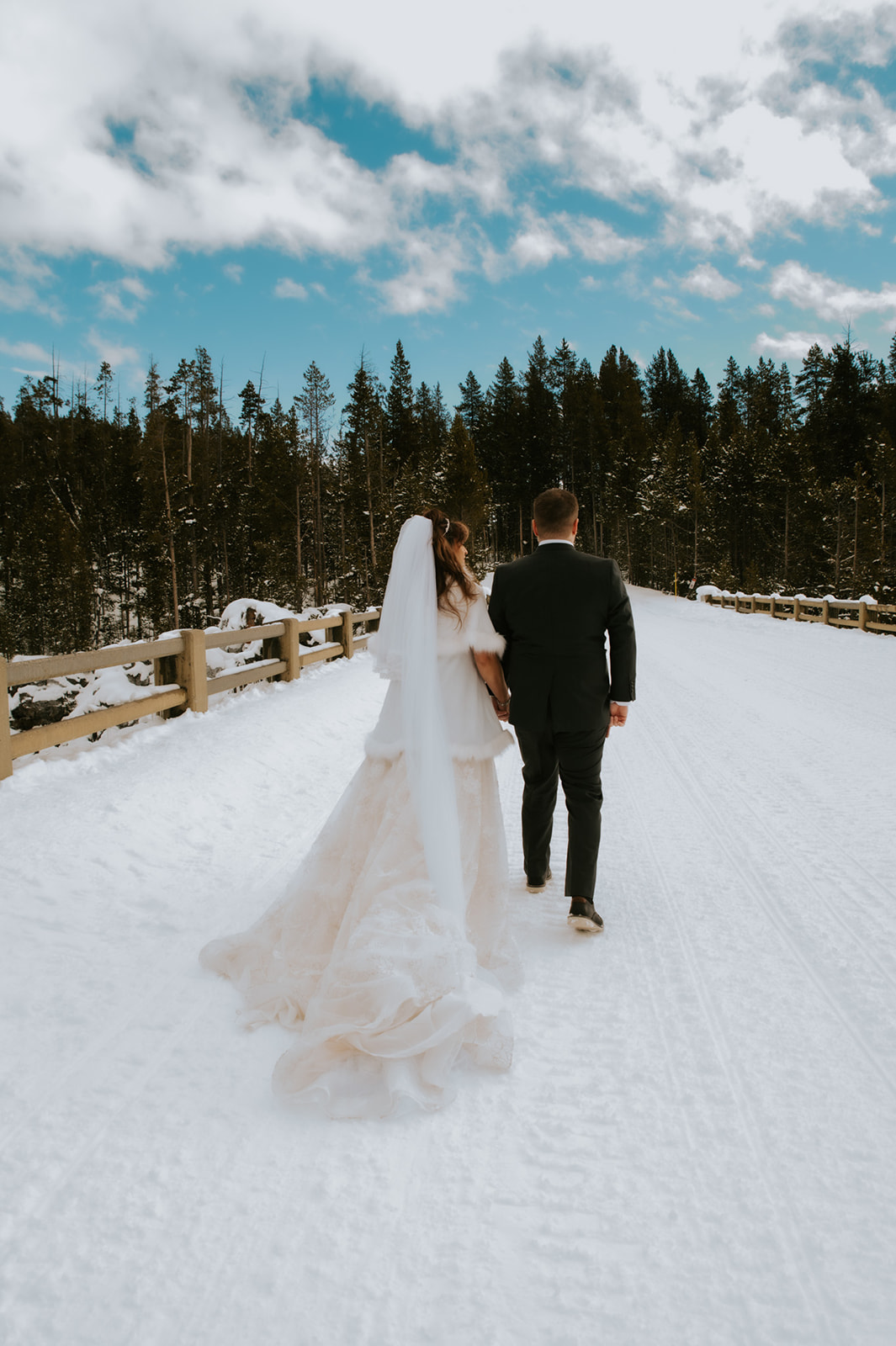 Couple walking away together on a snow-covered bridge beneath a blue sky, bride’s veil flowing behind her.