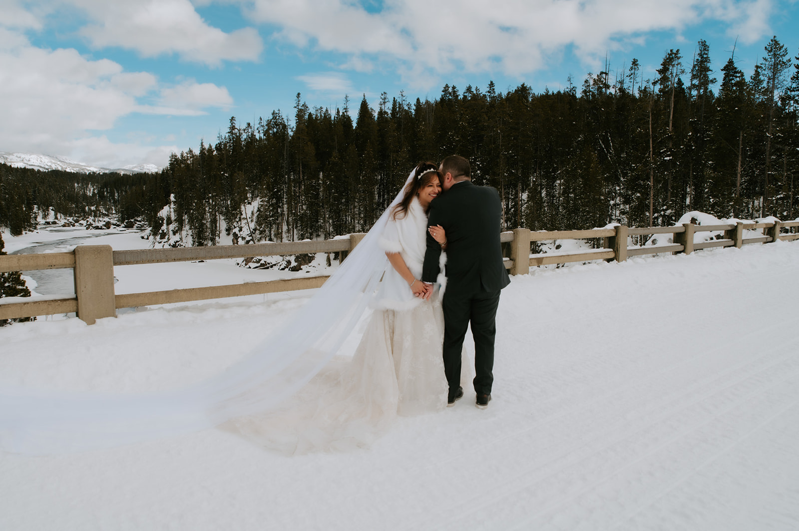 Bride smiling up at her groom as they stand hand in hand on a snow-covered bridge overlooking a frozen river and forest.