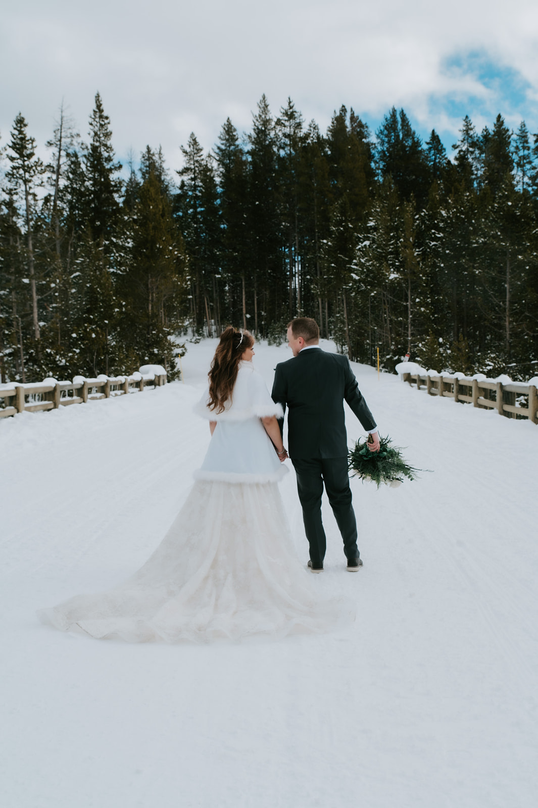 Newlywed couple walking hand in hand down a snow-covered forest road, bride’s long train trailing behind her.