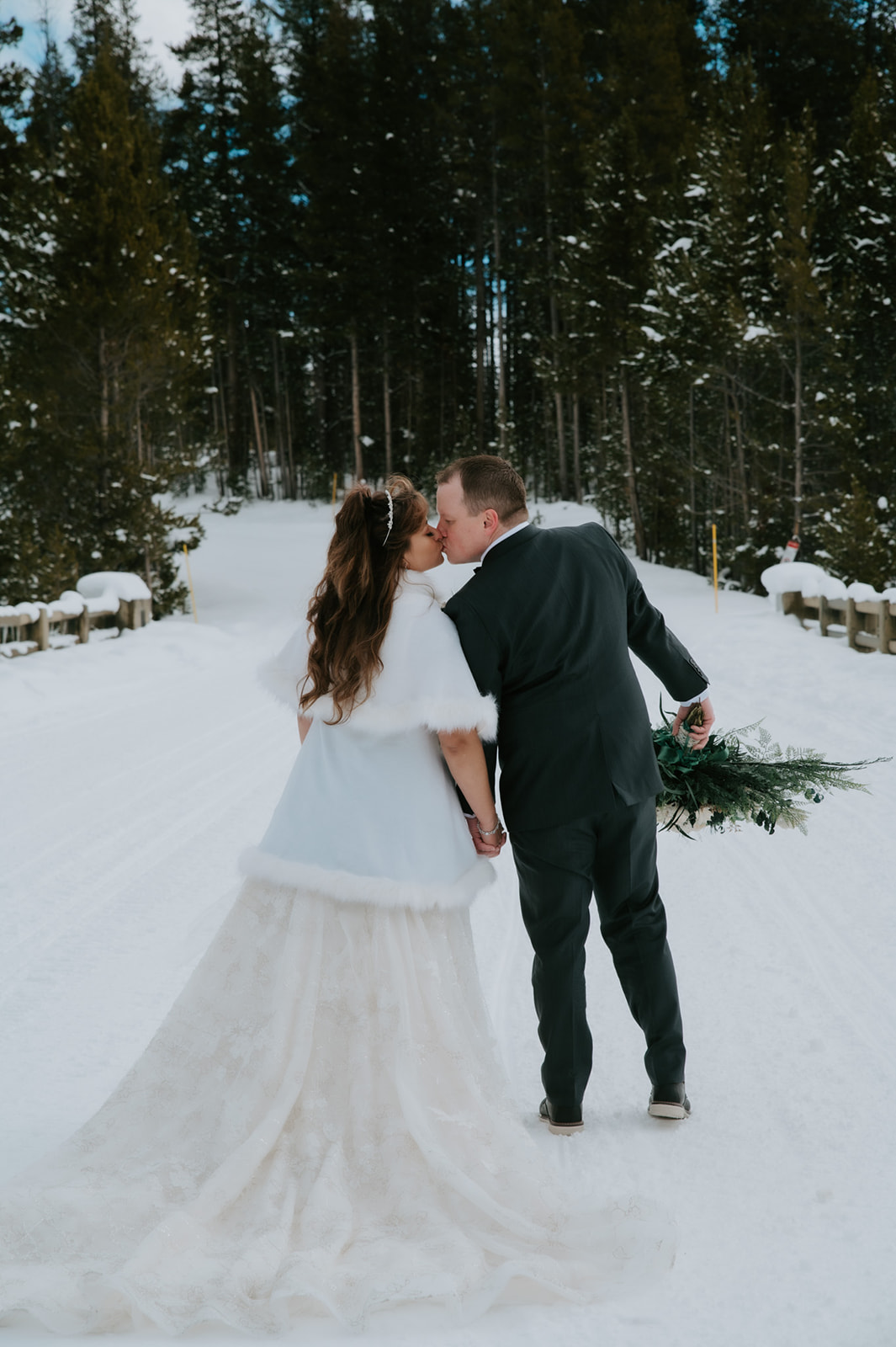 Bride and groom walking hand in hand down a snow-covered forest road, captured from behind during their romantic elopement video.