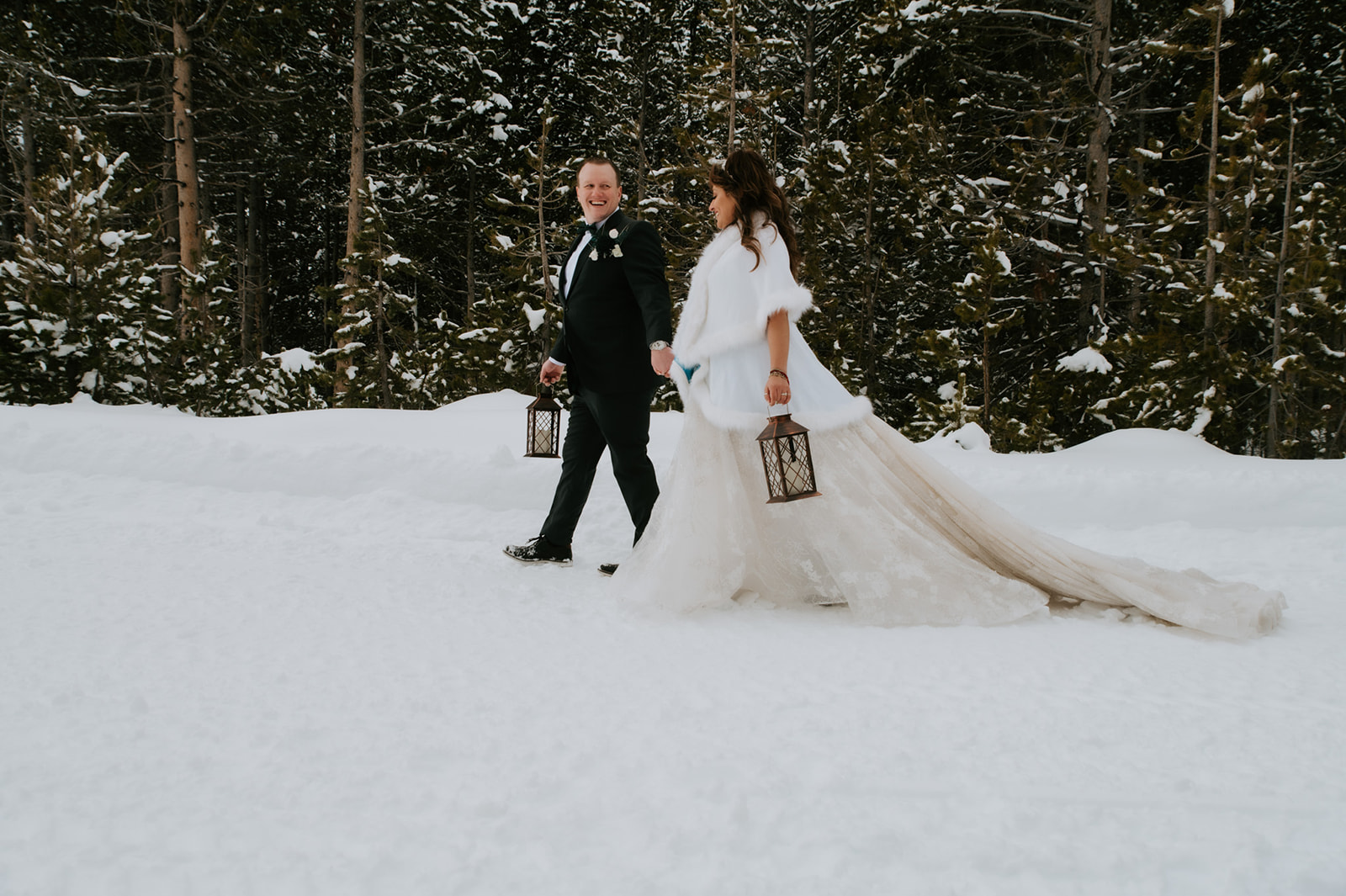 Bride laughing as she walks beside her groom through fresh snow, carrying lanterns beneath tall pine trees.