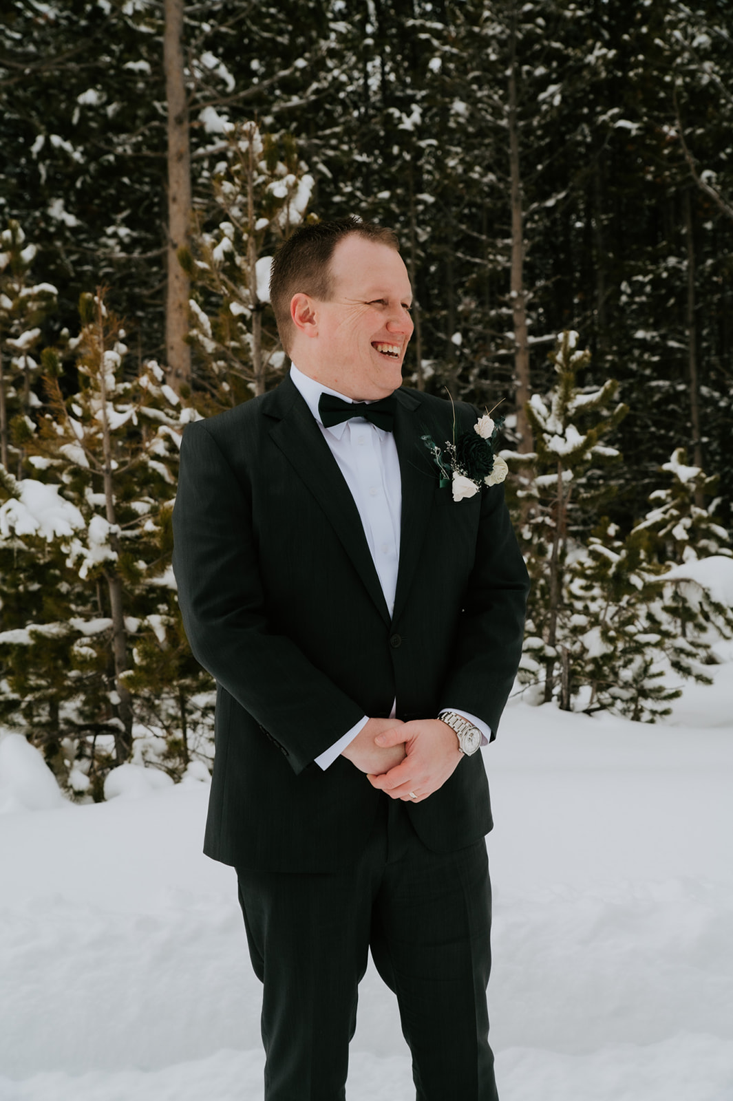 Groom smiling and waiting in the snow before the ceremony, surrounded by evergreen trees.