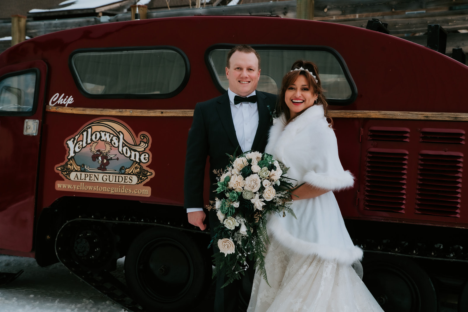 Bride and groom smiling in front of a vintage red snowcat in Yellowstone, holding a cascading bouquet of white and deep green roses.