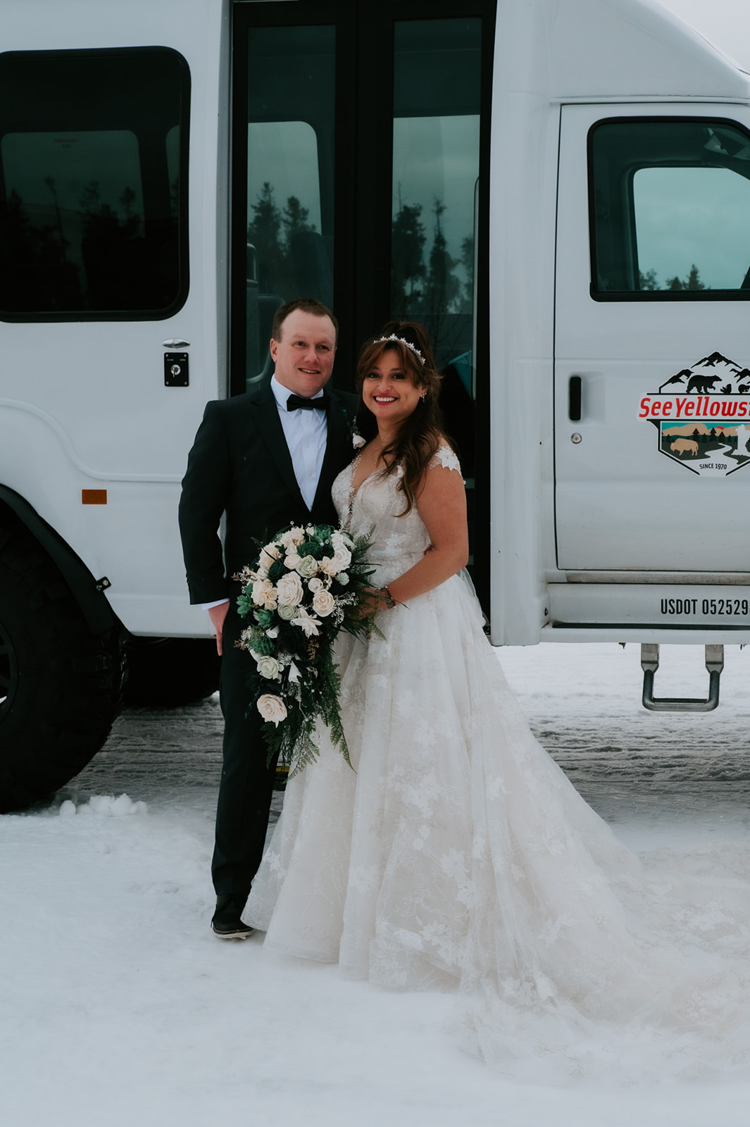 Bride and groom smiling in front of a white Yellowstone shuttle bus in the snow after their winter elopement video adventure.