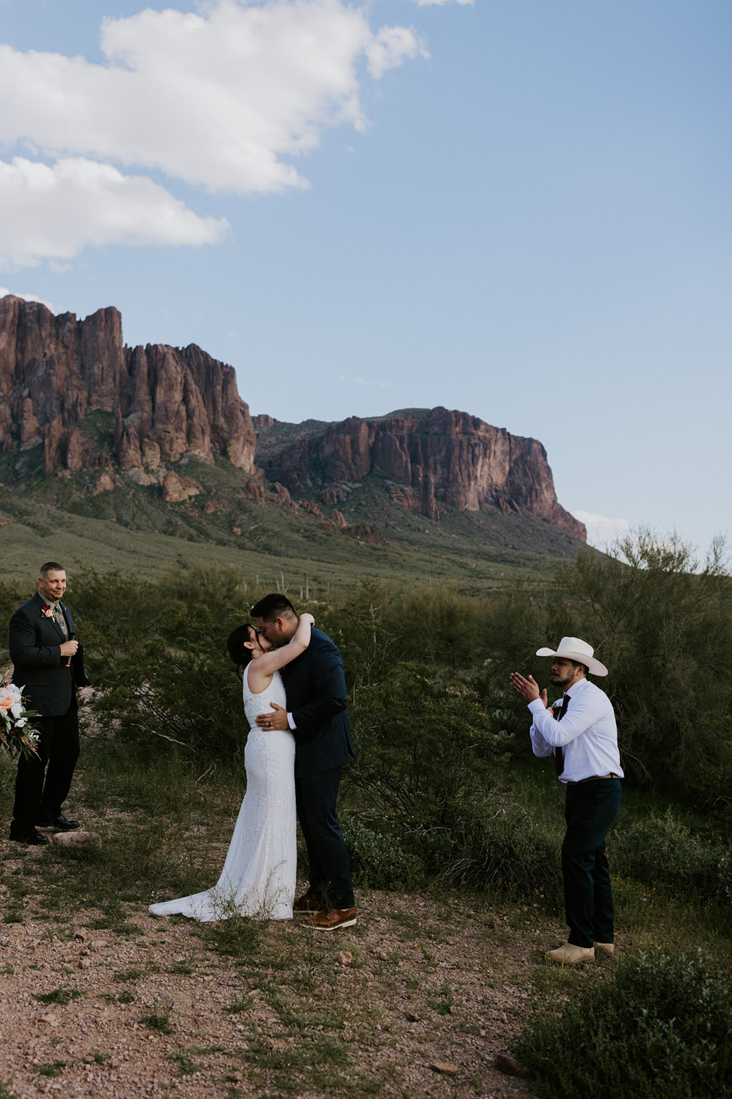 Bride and groom sharing their first kiss after vows as guests celebrate against a desert mountain backdrop