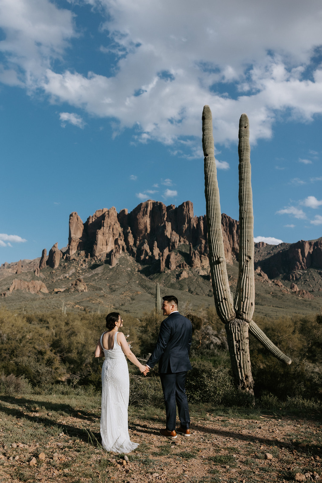 Arizona elopement portrait beneath tall saguaro cactus with red rock mountains and scattered clouds overhead