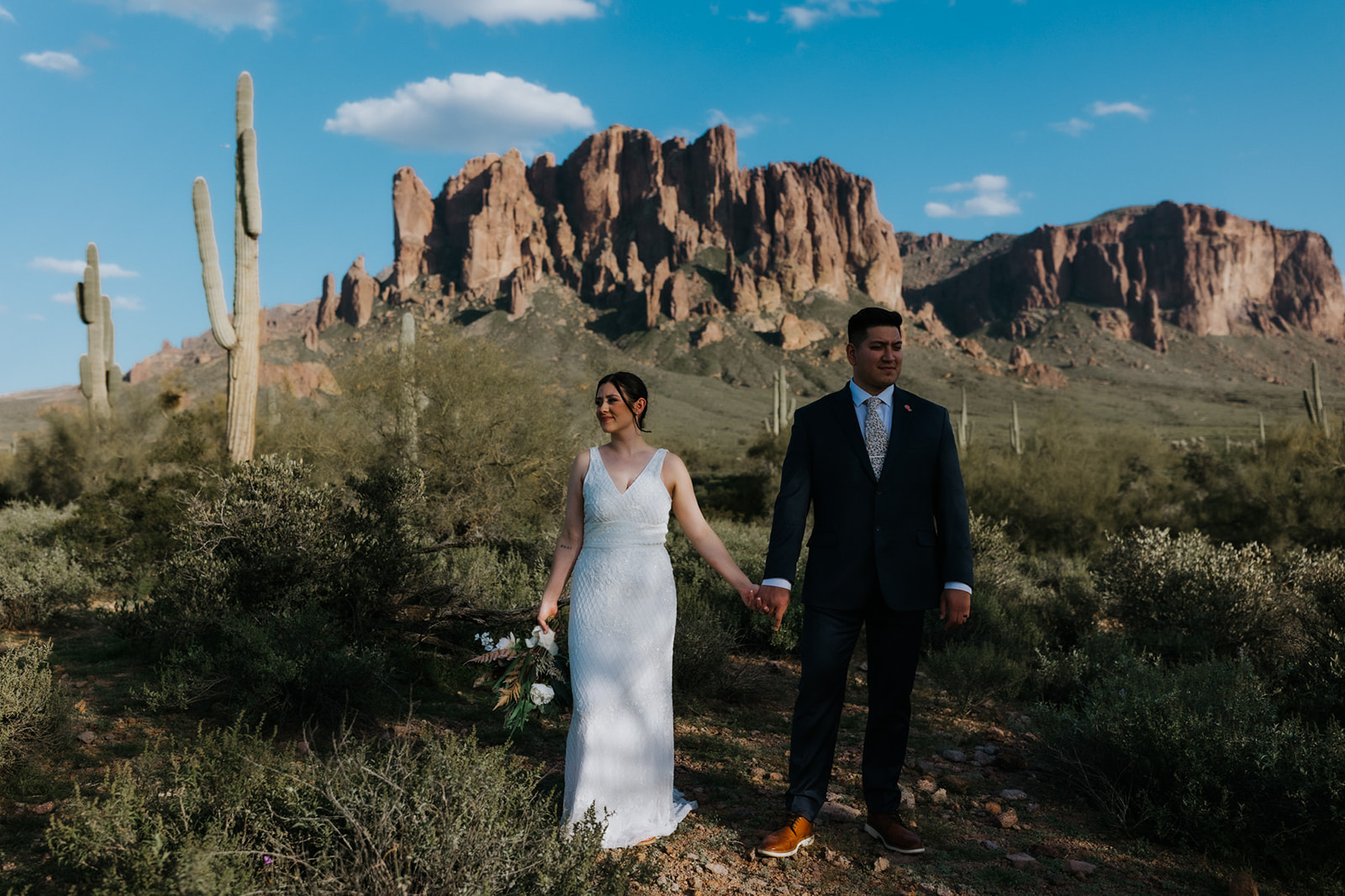 Bride and groom walking hand in hand through the desert with rugged mountain backdrop and blue sky
