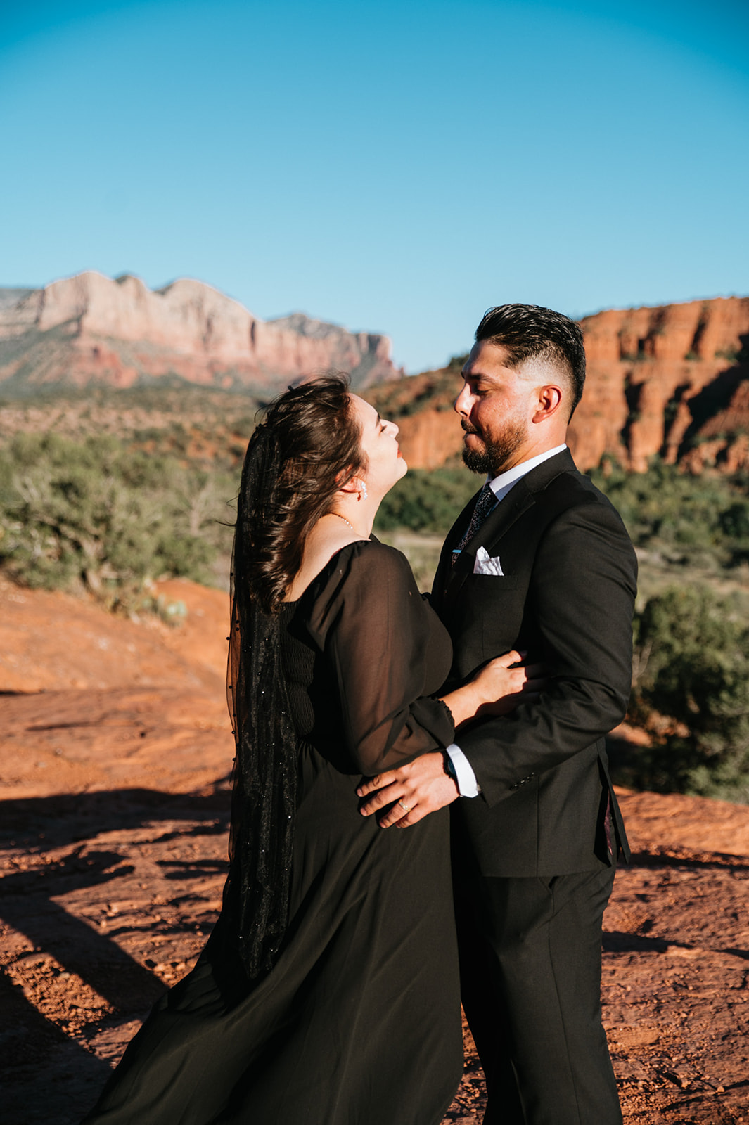 Bride in flowing black dress leaning into groom on red rock ledge with Sedona buttes glowing behind them