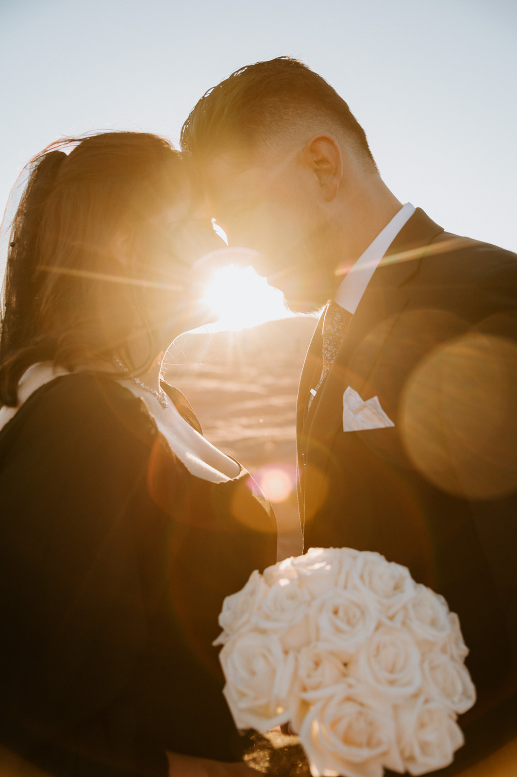 Backlit Arizona elopement portrait with sun flare as couple leans in for a kiss, bride holding white rose bouquet