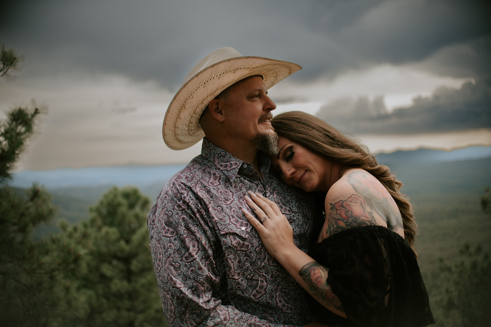 Close-up of tattooed bride resting her head on groom’s chest against a stormy mountain backdrop