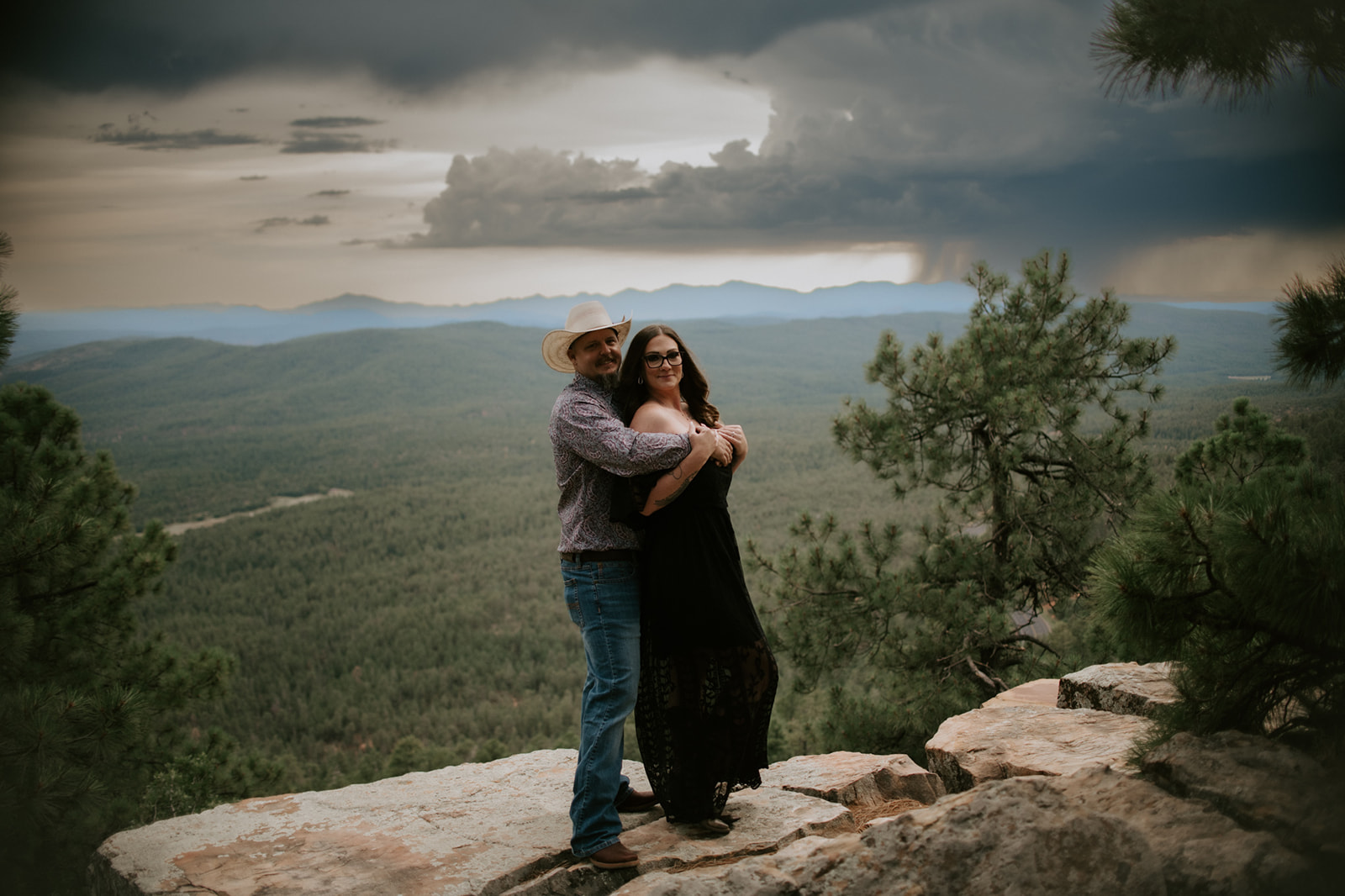 Couple standing together on rocky ledge with sweeping mountain views and dramatic storm sky