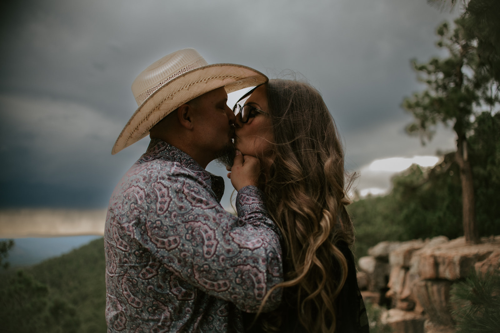 Intimate kiss beneath stormy skies on a cliffside overlook with pine trees and distant mountain views