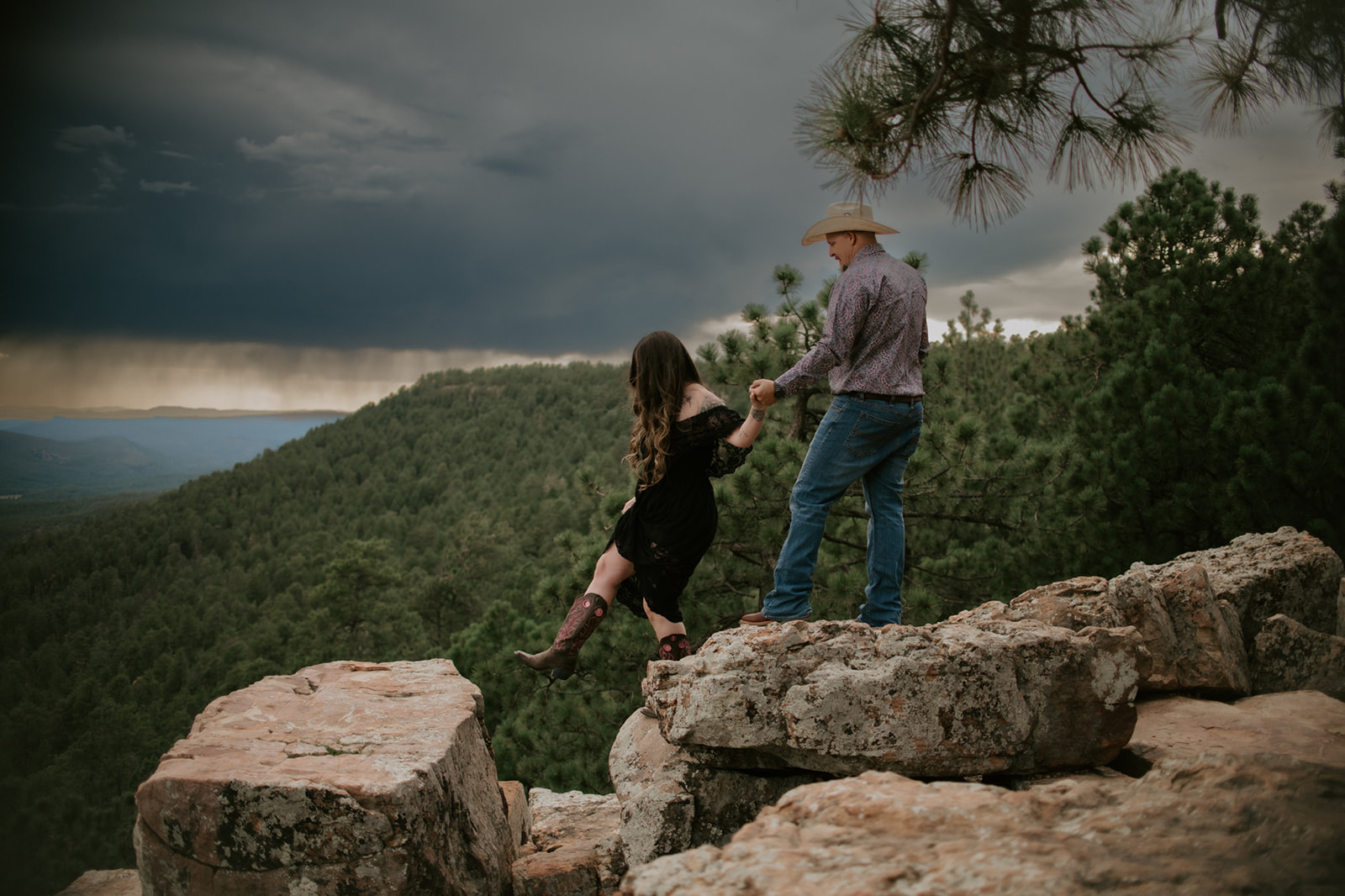 Couple holding hands while stepping across rocky cliff ledges under dramatic storm clouds in northern Arizona