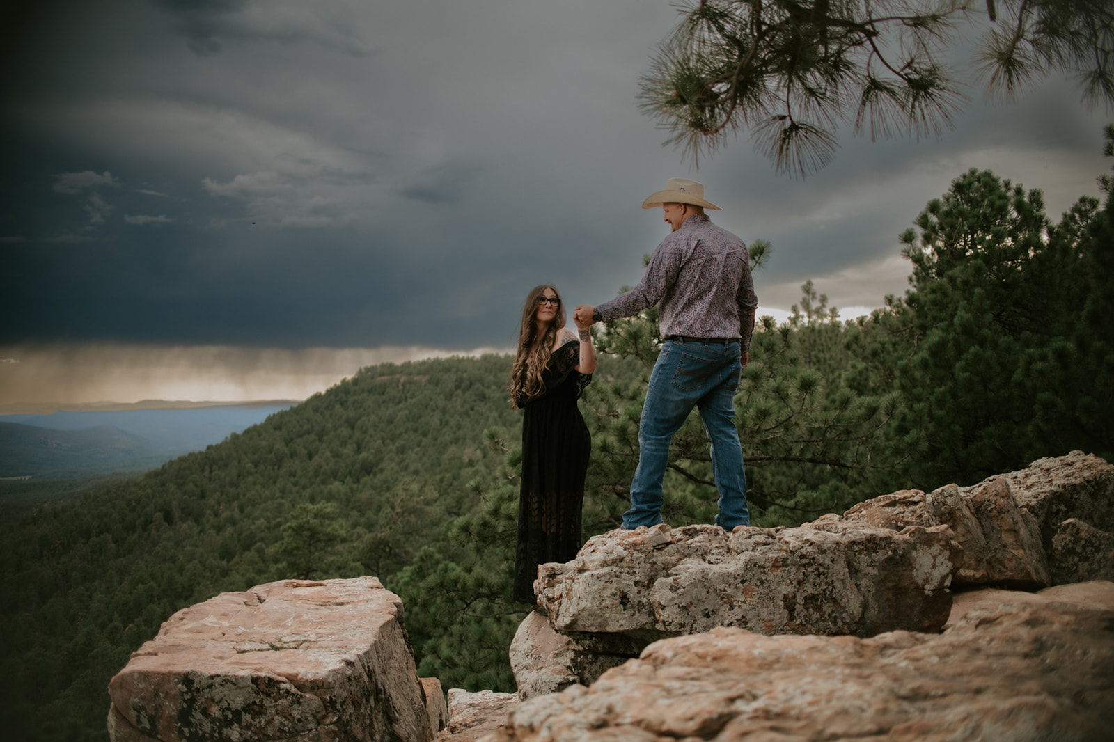 Couple holding hands on a rocky cliff under dramatic storm clouds and pine forest views in northern Arizona