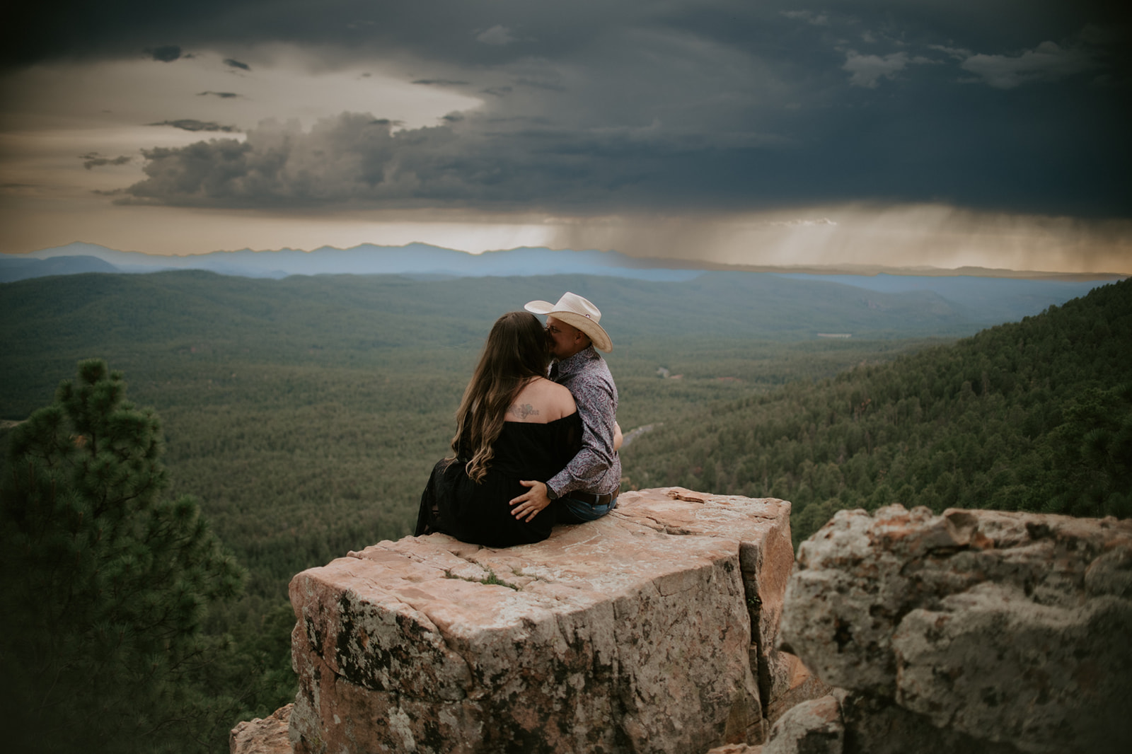 Couple sitting together on a cliff edge as rain falls in the distance over layered mountain ridges