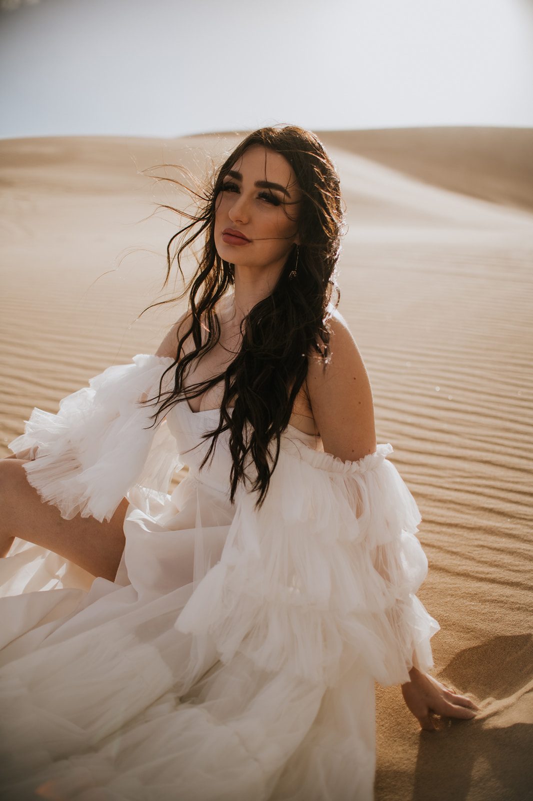 Bride sitting in soft desert sand with wind moving through her dark hair and ruffled elopement dress.
