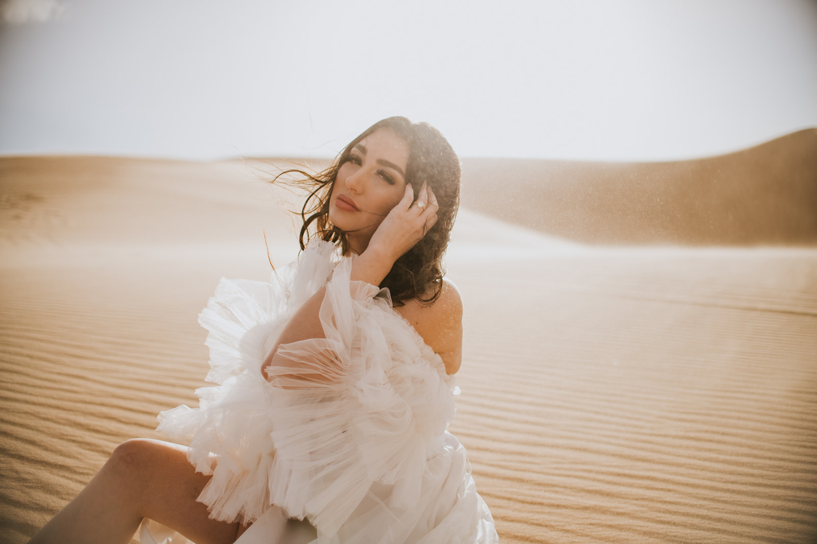 Bride sitting in rippled desert sand adjusting her hair as sunlight glows through the dunes.
