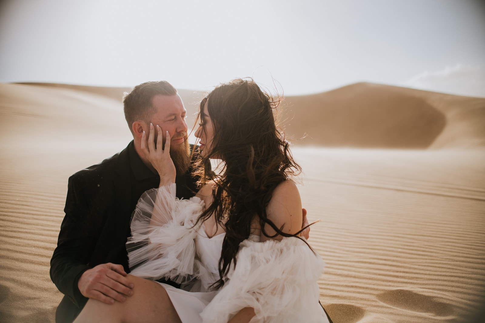 Bride gently touching the groom’s face as they sit together in sunlit desert dunes.
