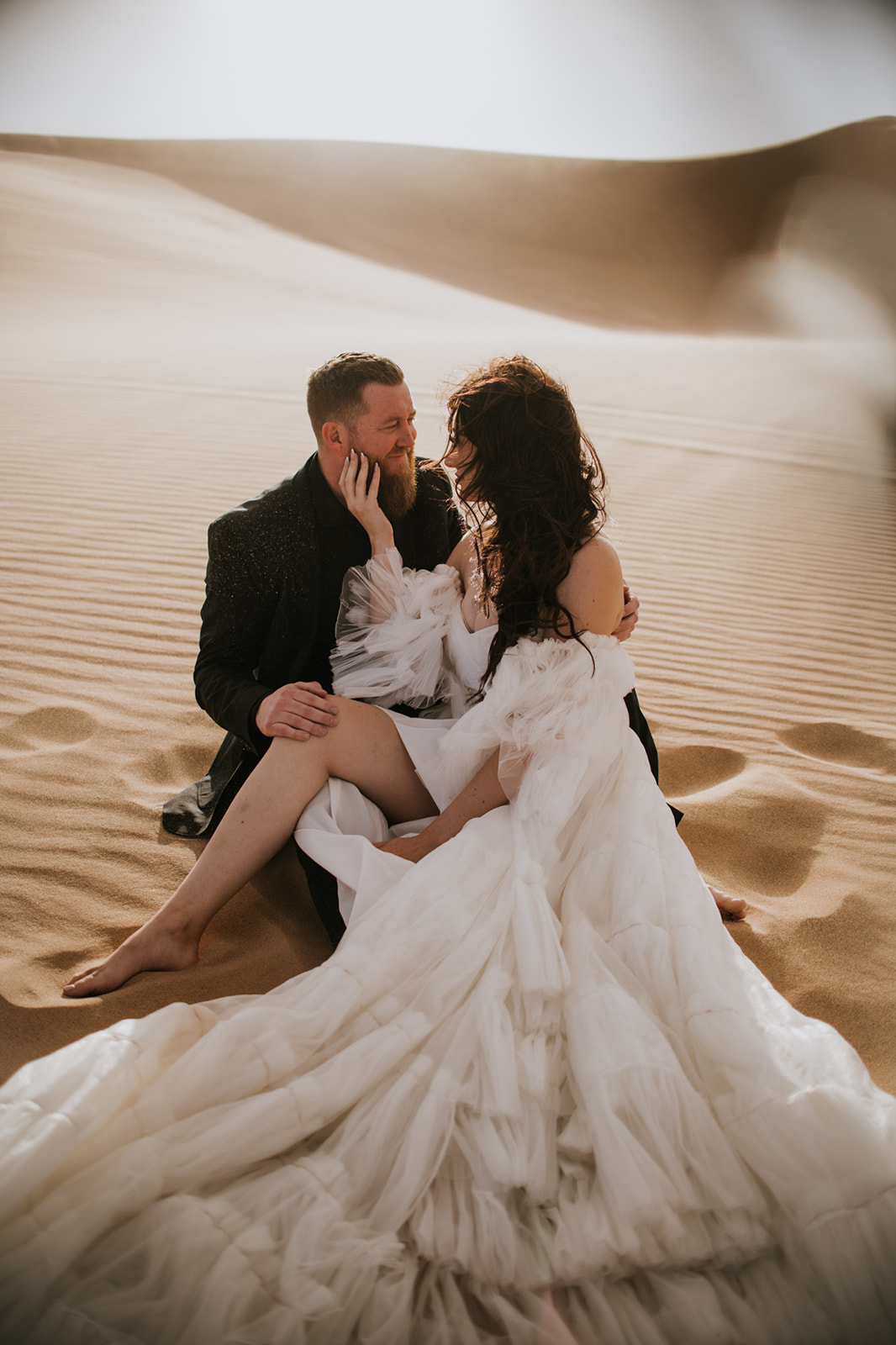 Bride and groom sitting close together in rippled sand dunes sharing an intimate moment during their destination elopement.
