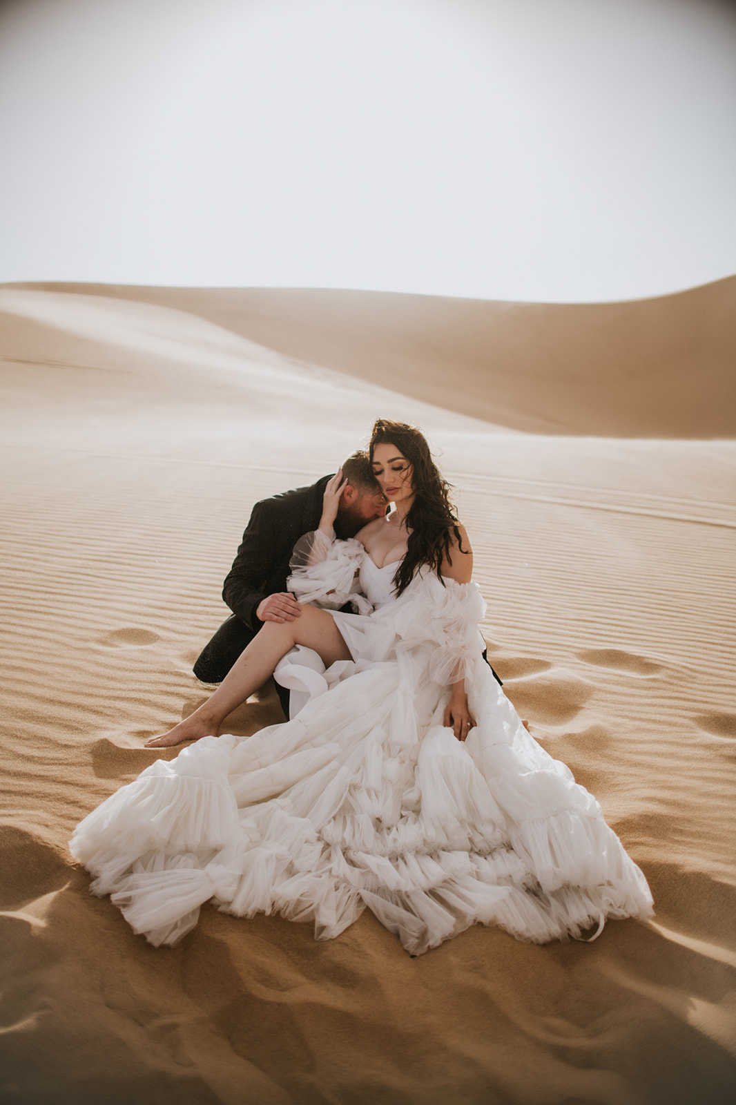 Bride in a flowing off-shoulder gown sitting in golden desert sand dunes while the groom leans close during a quiet destination elopement moment.
