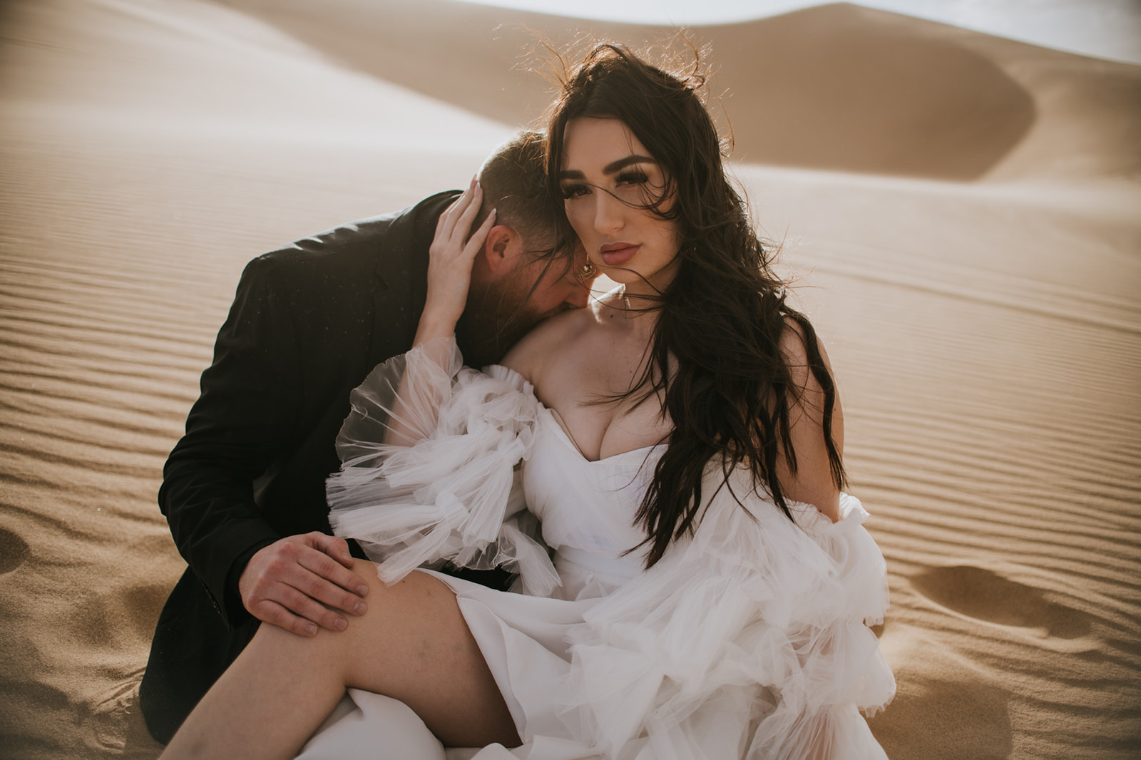 Bride sitting in rippled desert sand while the groom leans against her shoulder in an intimate moment.
