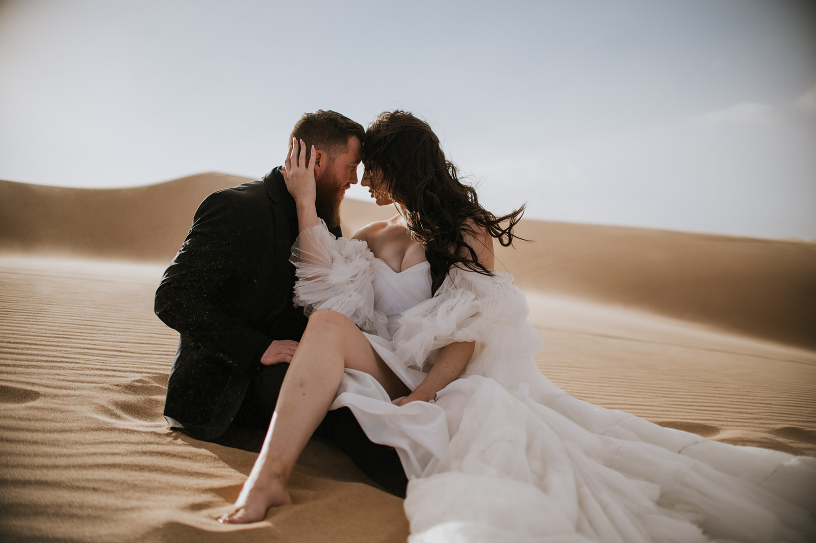 Couple sitting close together in the sand with wind moving through the bride’s flowing dress during their destination elopement.
