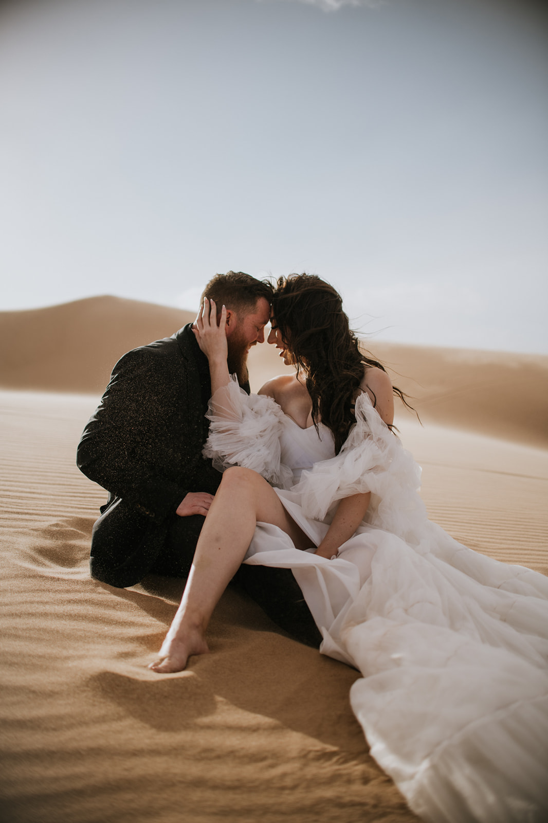 Couple sitting close together in soft desert sand dunes, foreheads touching in an intimate moment.
