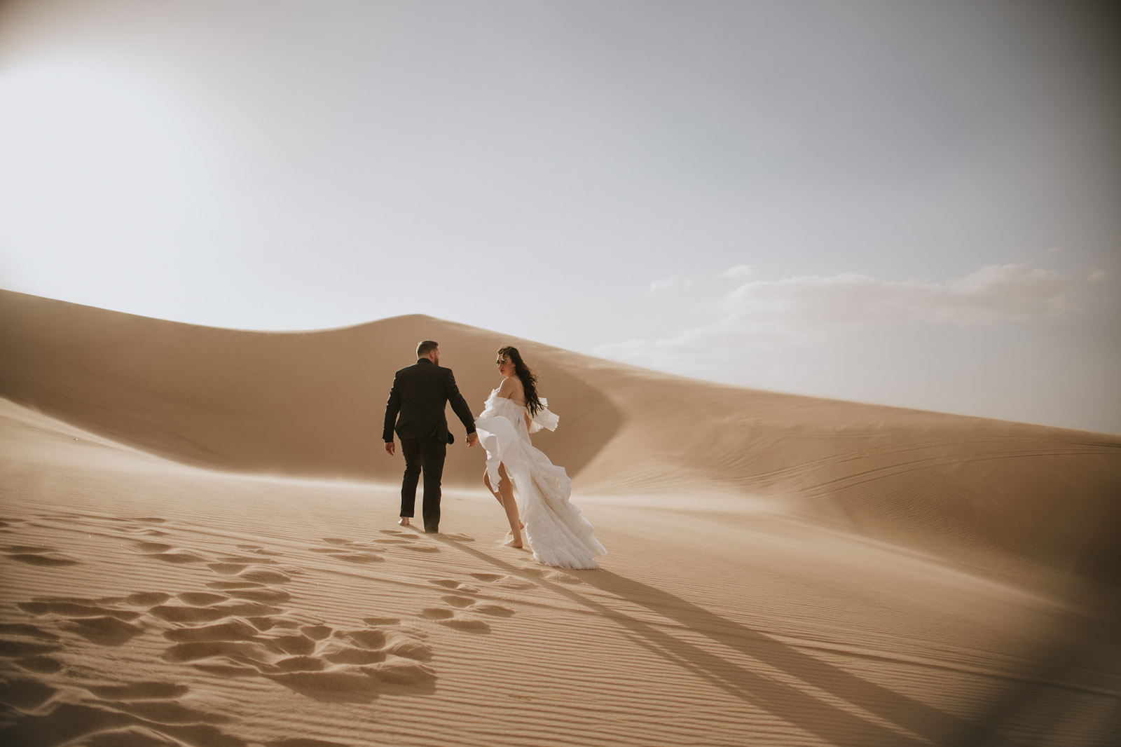 Newlywed couple walking hand in hand across sweeping desert dunes at golden hour.
