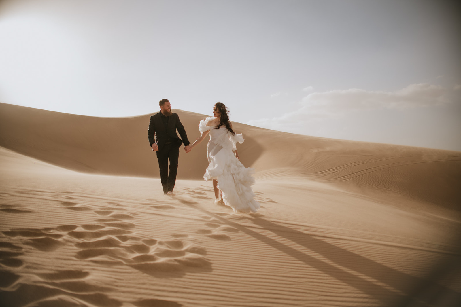 Adventurous couple running across expansive desert sand dunes with long shadows stretching behind them.
