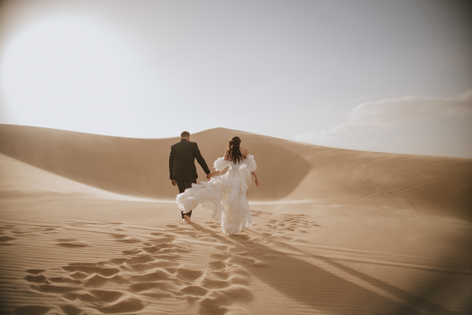 Newlywed couple running hand in hand across wide desert dunes during an adventurous destination elopement.
